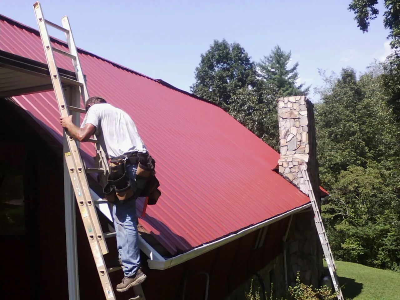 Roofer on a ladder beside a red metal roof and stone chimney — Soffit & Fascia Repair by Roofing Brothers in Rebuck